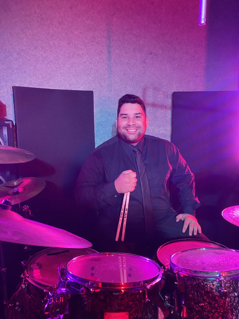 Eduardo Pirajá sentado à bateria com baquetas, em show com iluminação cênica